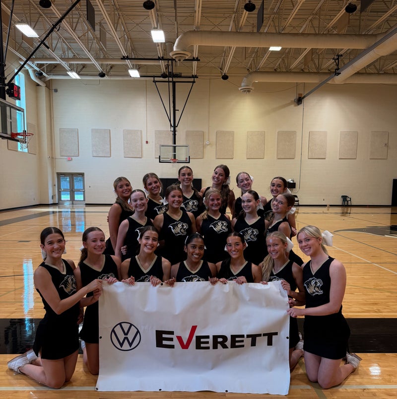 A large group of smiling cheerleaders in black and gold uniforms kneeling and holding a banner in a gym