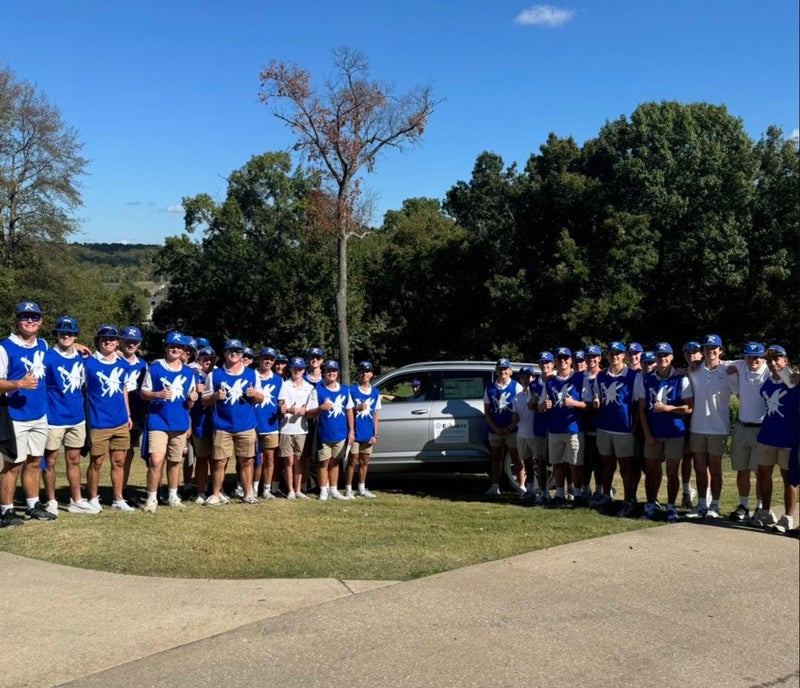 A large group of male students in matching blue shirts, golf hats, and khaki shorts pose thumbs-up on a golf course