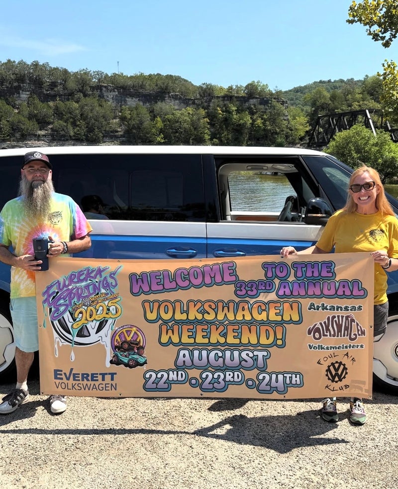 Two people hold a colorful banner announcing the 33rd Annual Volkswagen Weekend! against a scenic backdrop