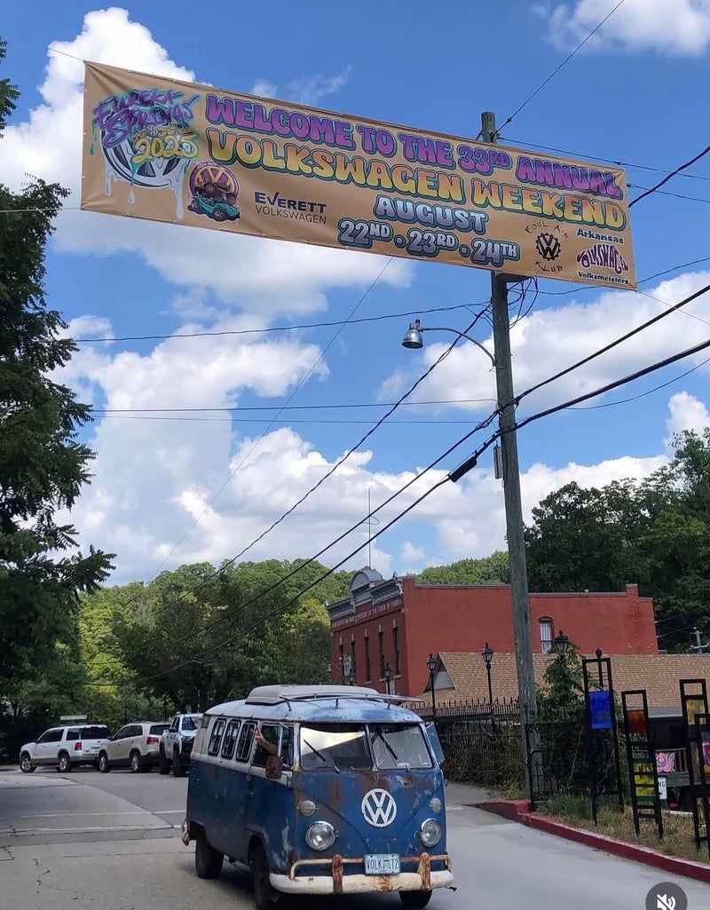 A rusty blue Volkswagen van drives under a banner for the 33rd Annual Volkswagen Weekend in Eureka Springs.