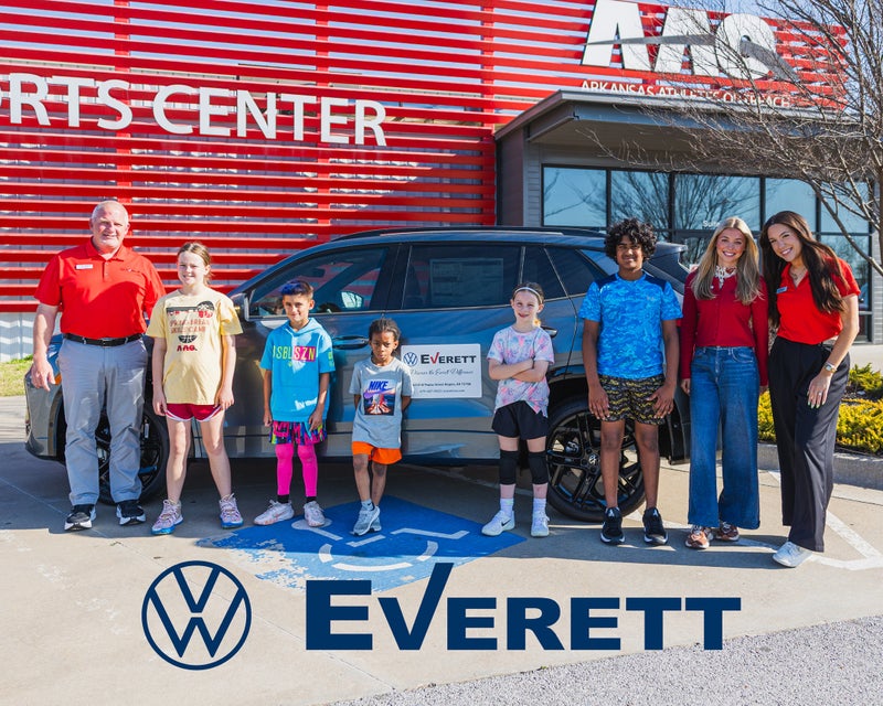 A man, two women, and five children pose with a black SUV in front of a building with a red slatted facade