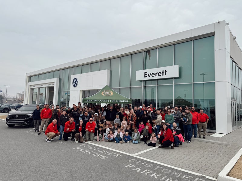 A large group of people including many children, pose in front of the Everett Volkswagen dealership under a cloudy sky.