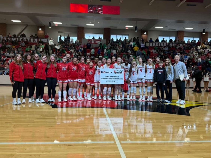 A girls' basketball team in red outfits and 2 male adults pose on a gym floor with a championship banner.