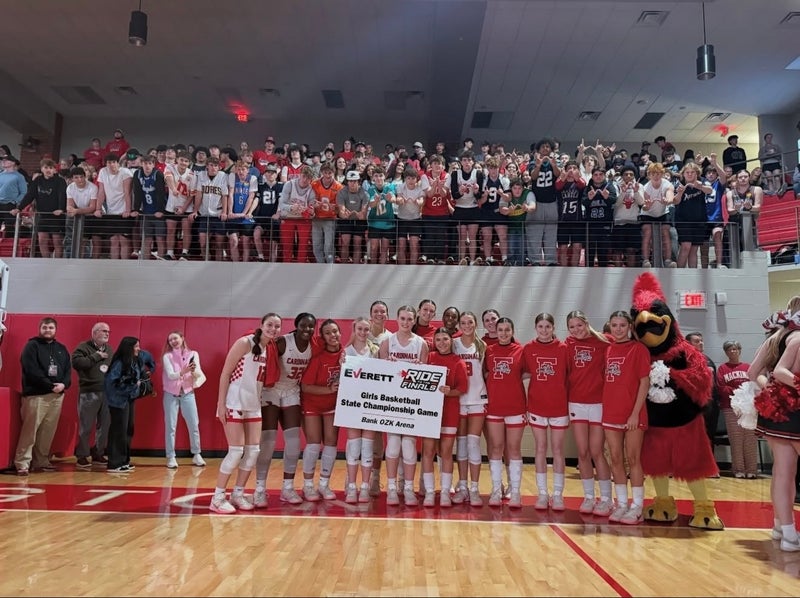A girls' basketball team in white and red uniforms poses with a State Championship Game sign, a mascot, and fans