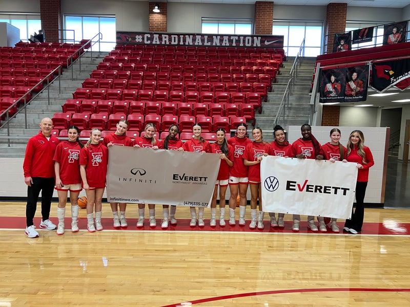 A women's basketball team and coaches hold sponsor banners in a gym.