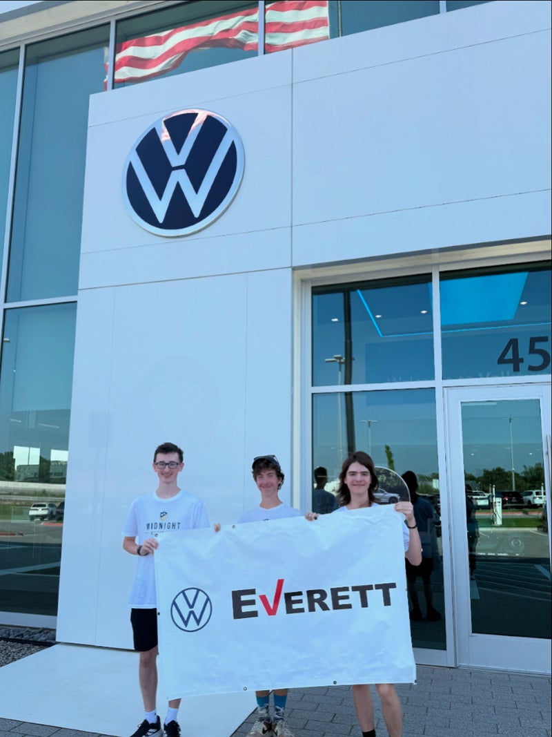 Three young men hold a white 'Everett' banner in front of a VW dealership with an American flag reflected above