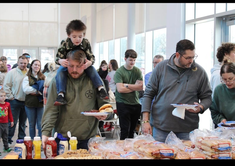 A man holding a plate of food with a child on his shoulders, in a crowded indoor cafeteria with condiments and bread.