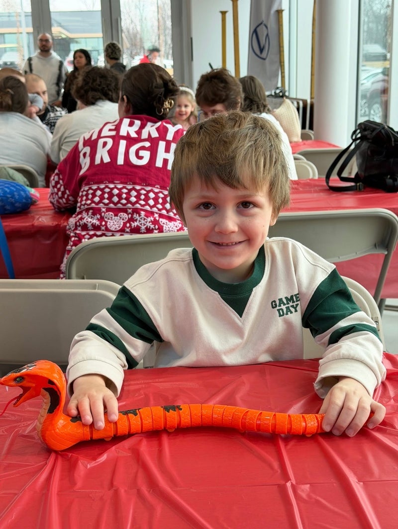 A young boy with short brown hair, wearing a Game Day shirt, holds an orange toy snake and smiles.