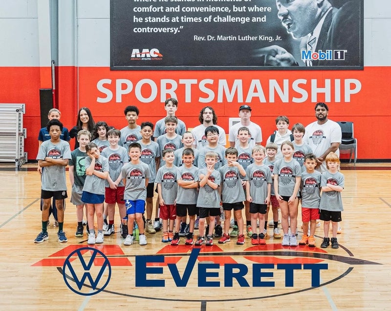 A large group of children and adults pose for a photo on a basketball court with SPORTSMANSHIP on the wall behind them.