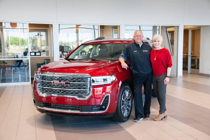 A smiling man and woman stand next to a shiny red GMC SUV inside a dealership showroom.